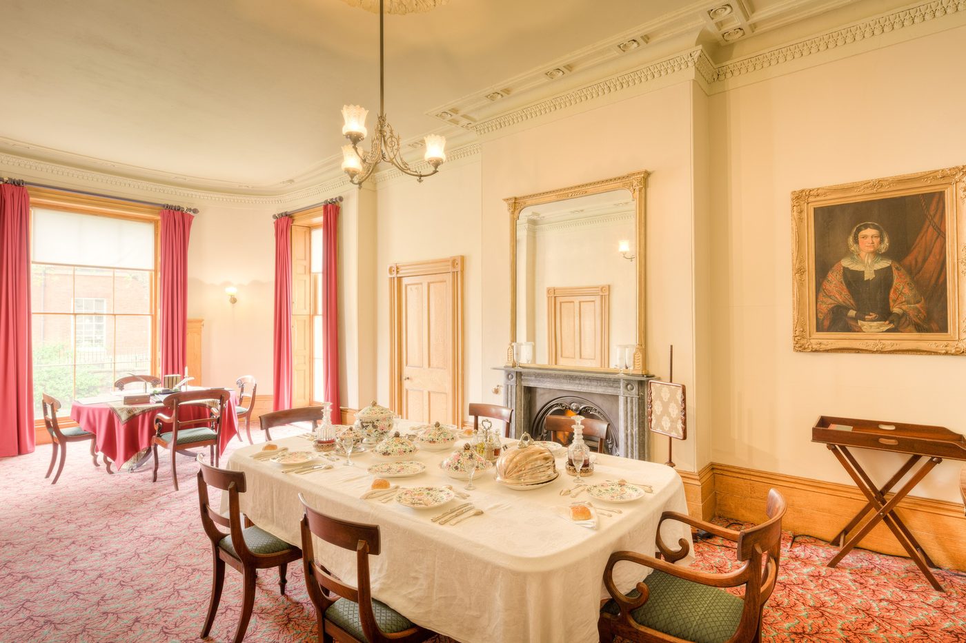 A dining room with a long laid table, a fireplace and tall windows hung with red curtains