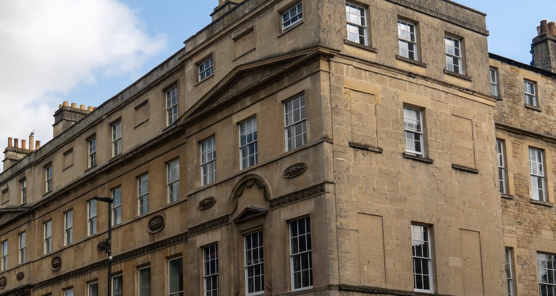 The corner of a Georgian terrace in Bath built of honey-coloured stone, with rows of tall sash windows and a pedimented doorcase