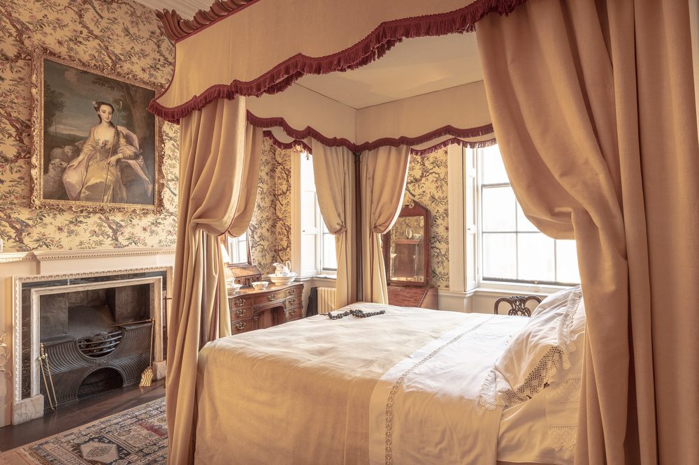 A Georgian bedroom with a curtained four-poster bed, a marble fireplace and a tall sash window with heavy drapes